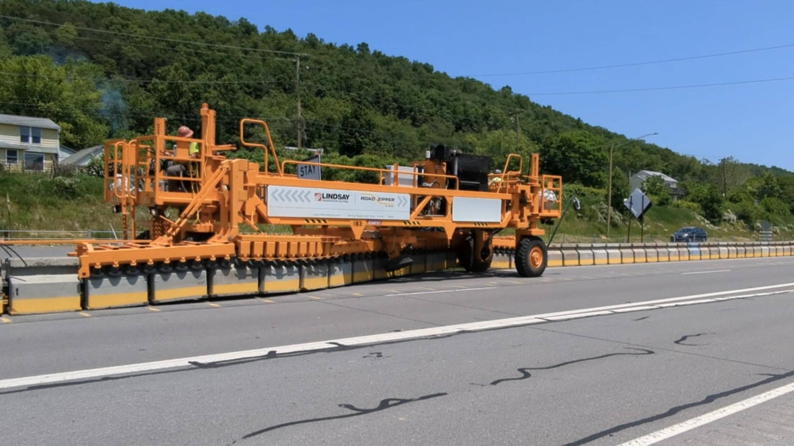 An image of a large orange piece of equipment being operated by two drivers in hard hats and yellow safety vests positioned on either end moving jersey barriers from one place on the roadway to another